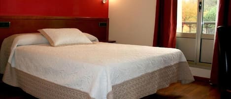 A bed with a white bedspread and two pillows, a wooden headboard, a nightstand, a window with red curtains, and a view of trees outside.