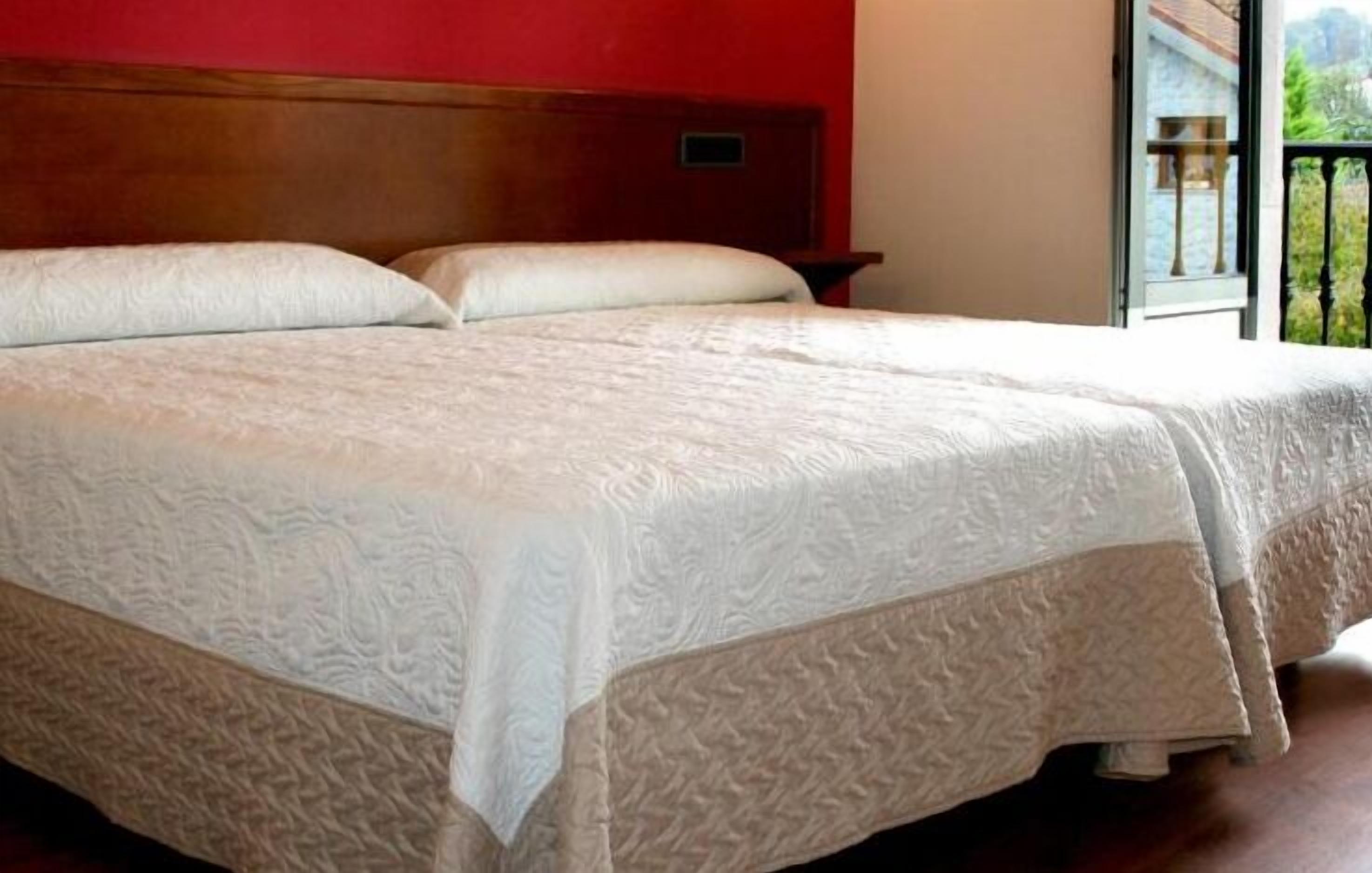 Cama doble con colcha blanca texturizada, cabecera de madera y vista a la vegetación a través de una ventana.