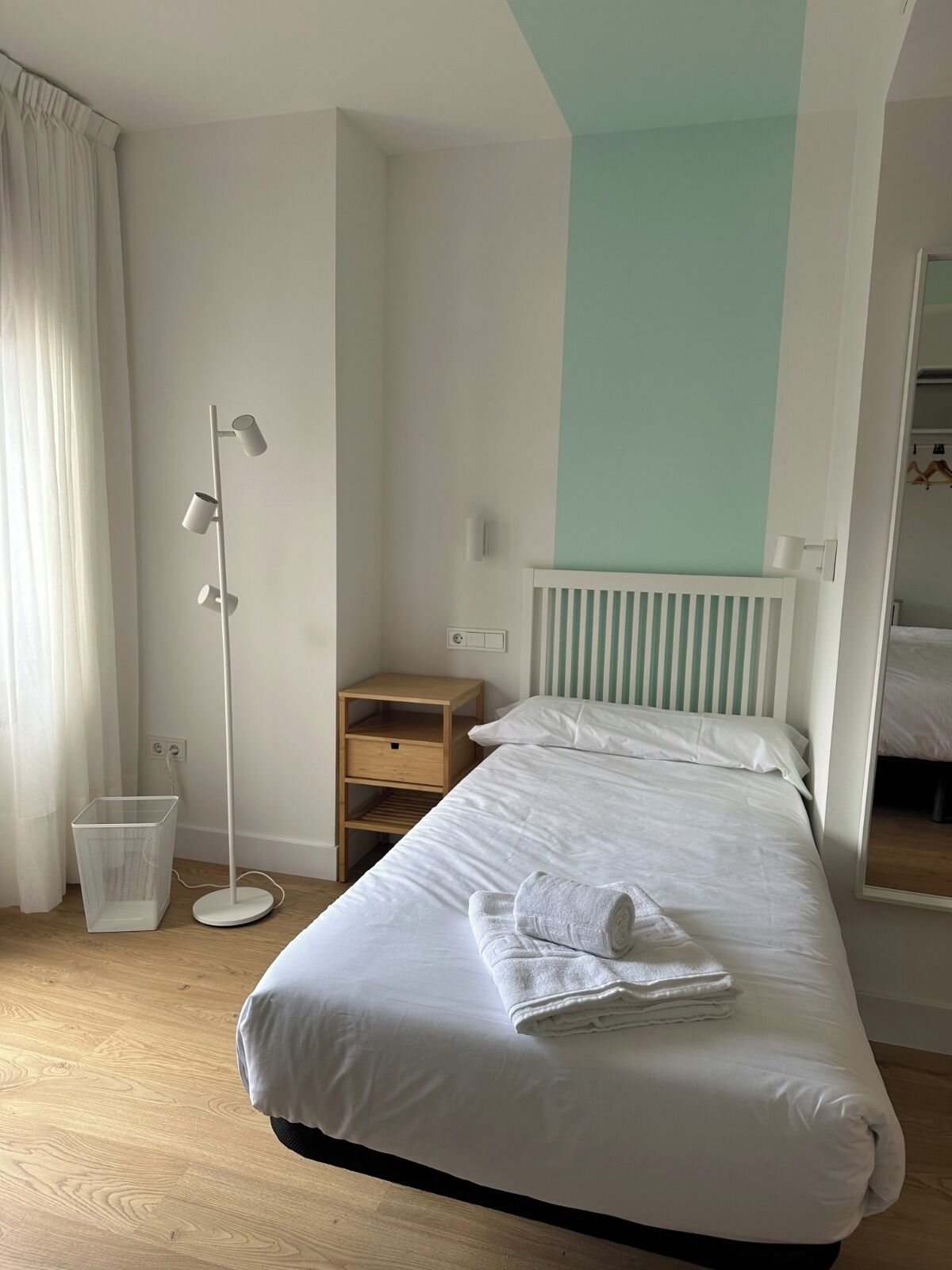 Una camera d'albergo con un letto rifatto, un comodino, una lampada da terra e un piccolo mobile contenitore.