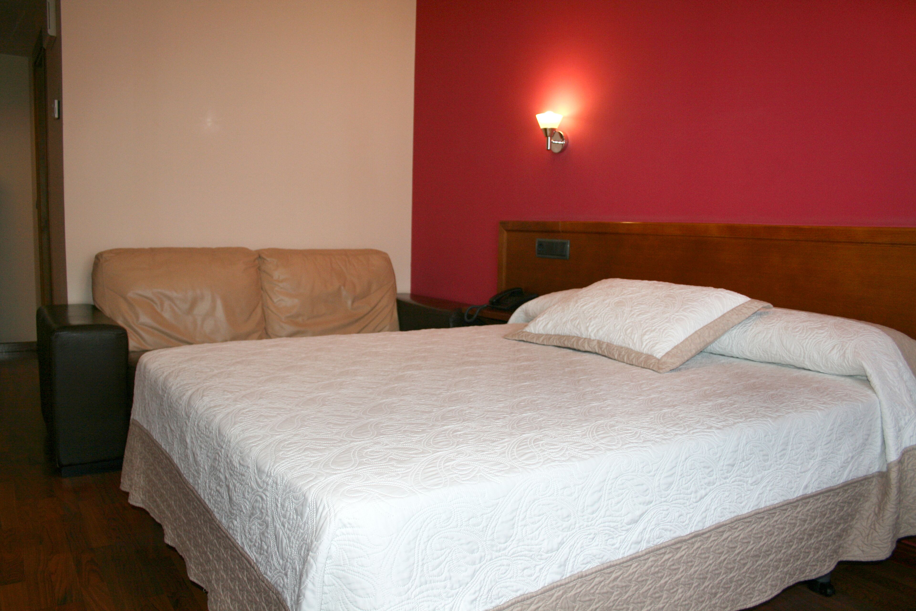 Una cama bien hecha con colcha blanca, cabecera de madera y pared roja con una lámpara de pared.