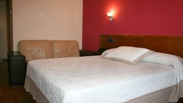 Una cama bien hecha con colcha blanca, cabecera de madera y pared roja con una lámpara de pared.