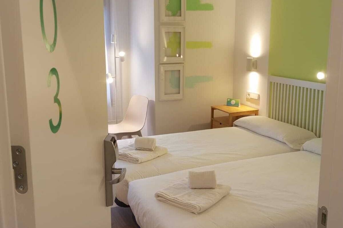 Habitación de hotel con dos camas, una silla, una mesita de noche y una pared con un número.