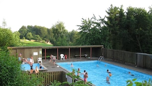 Ein Schwimmbeckenbereich mit überdachter Terrasse, Holzzaun und Rasenfläche im Hintergrund.