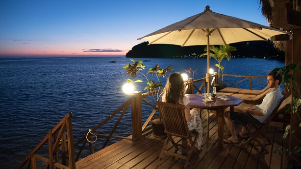 A wooden deck with a table and chairs, overlooking a body of water at dusk. - Aviavy Plage Hôtel (Nosy Be)