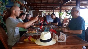 A wooden table with a menu, a straw hat, and a group of people dining. - Aviavy Plage Hôtel (Nosy Be)