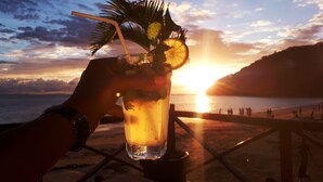 A hand holding a glass with a drink, palm leaves, and a straw, against a sunset background with a beach and people in the distance. - Aviavy Plage Hôtel (Nosy Be)