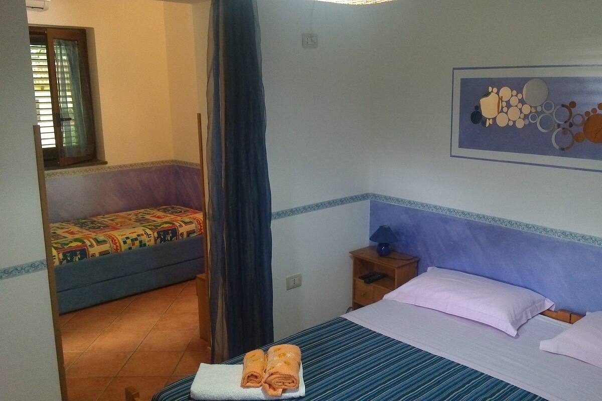 Une chambre d’hôtel avec un lit, une petite table, une table de chevet, une fenêtre avec des stores et un tableau au mur.