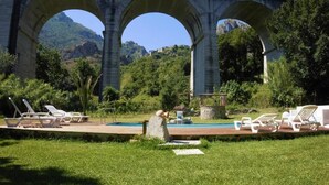 A pool area with lounge chairs and a fountain, under a large stone arch bridge with a mountainous backdrop. - B&B Donna Maja (Centola)