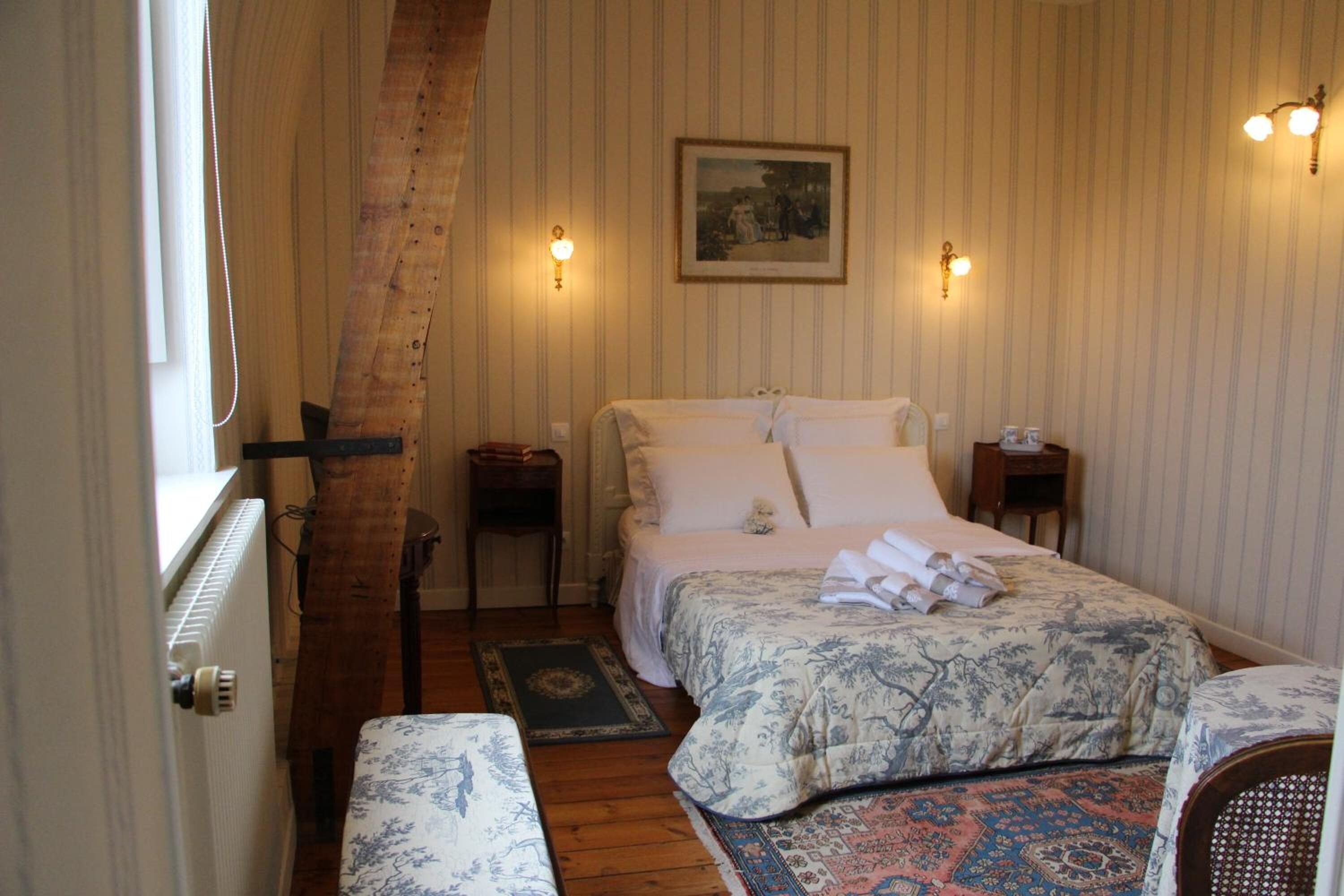 Une chambre avec un plafond en pente en bois, un grand lit recouvert d’une couvre-lit à motifs floraux, deux tables de chevet avec des lampes, un petit tapis et une fenêtre avec un rideau.