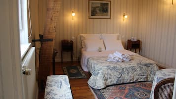 Une chambre avec un plafond en pente en bois, un grand lit recouvert d’une couvre-lit à motifs floraux, deux tables de chevet avec des lampes, un petit tapis et une fenêtre avec un rideau.