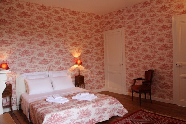 Un dormitorio con papel tapiz floral, una cama con ropa blanca, dos lámparas de noche, una silla de madera y una puerta.