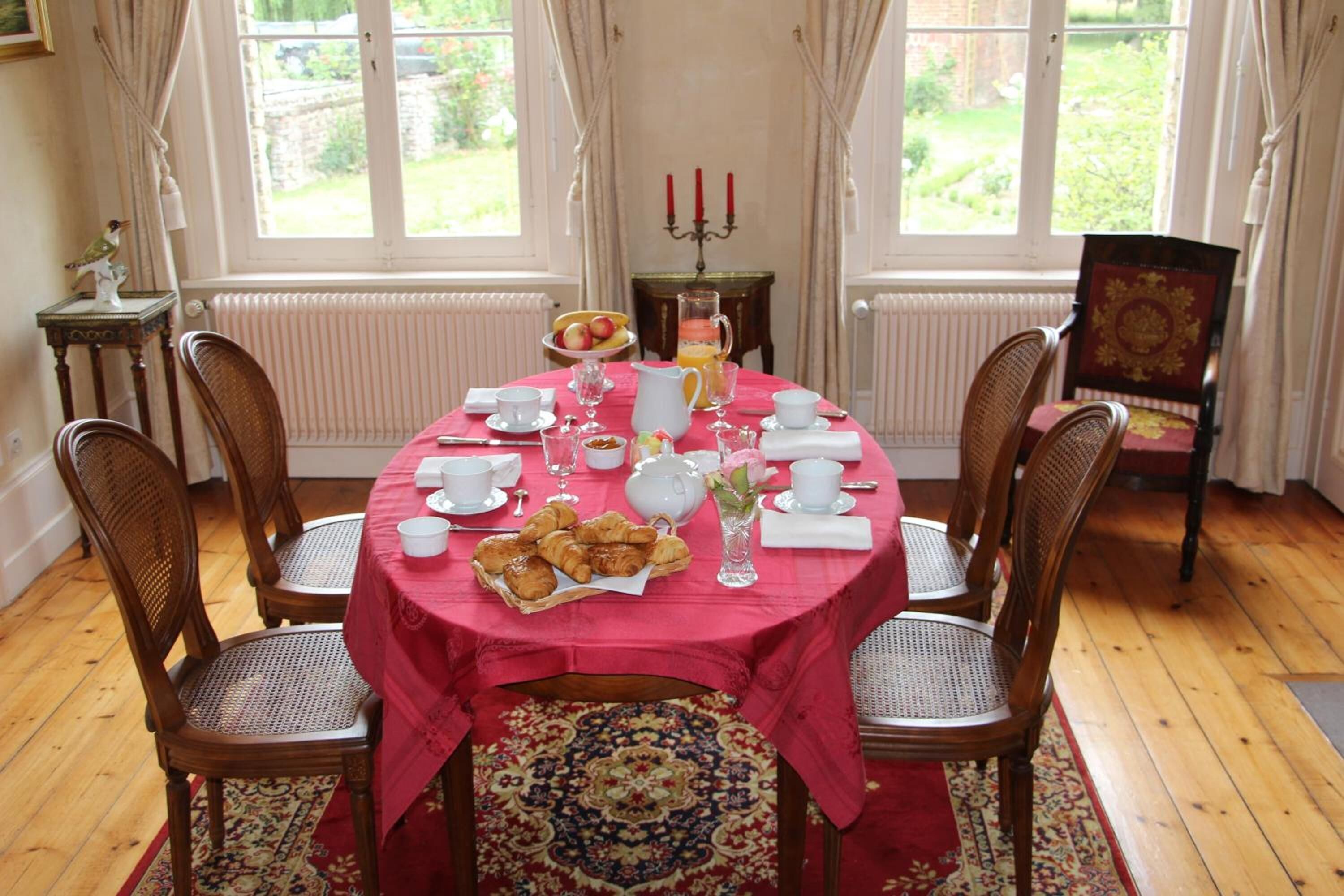A dining room with a round table set for six, covered with a red tablecloth, surrounded by wooden chairs.