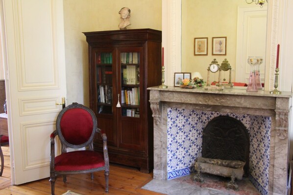 Una habitación con una estantería de madera, un sillón tapizado en rojo, una chimenea con azulejos de diseño azulado y una puerta con panel decorativo.