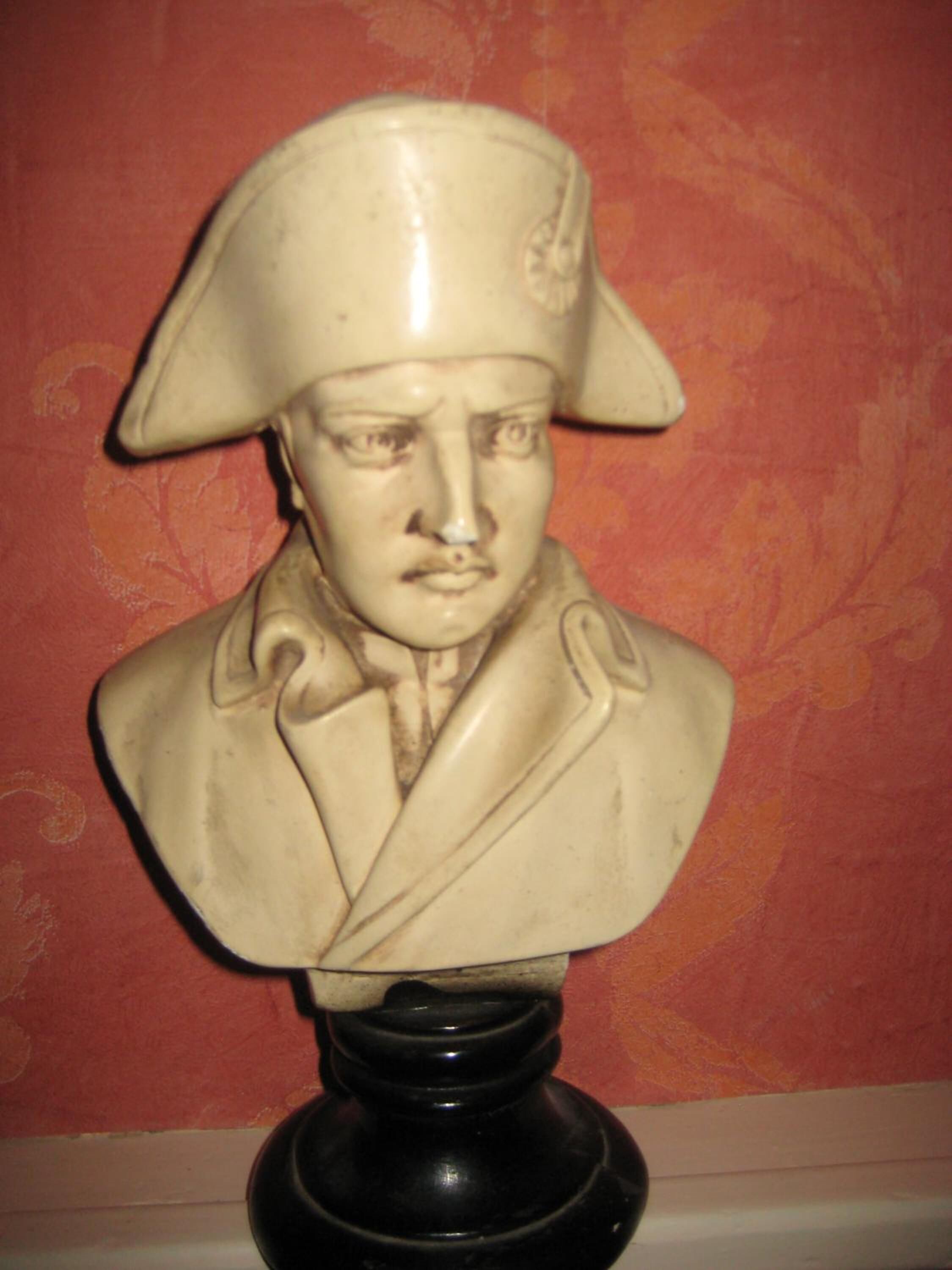 Una escultura de busto sobre un pedestal negro frente a una pared roja con diseño.