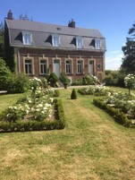 Uma grande casa de tijolos com dois andares, fachada simétrica, várias janelas e um jardim bem cuidado com flores brancas e sebes.