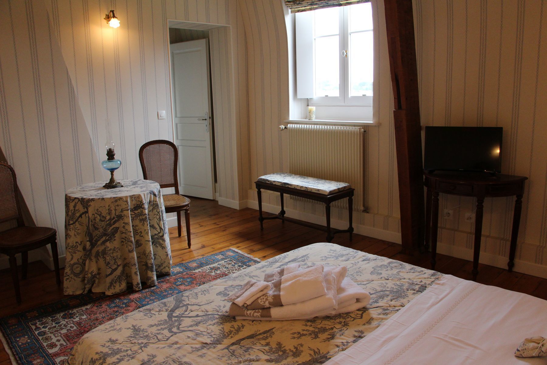 Un dormitorio con cama, una silla, una mesita con lámpara, un banco, un televisor sobre un mueble bajo y una ventana con vistas al exterior.