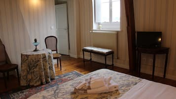 Un dormitorio con cama, una silla, una mesita con lámpara, un banco, un televisor sobre un mueble bajo y una ventana con vistas al exterior.