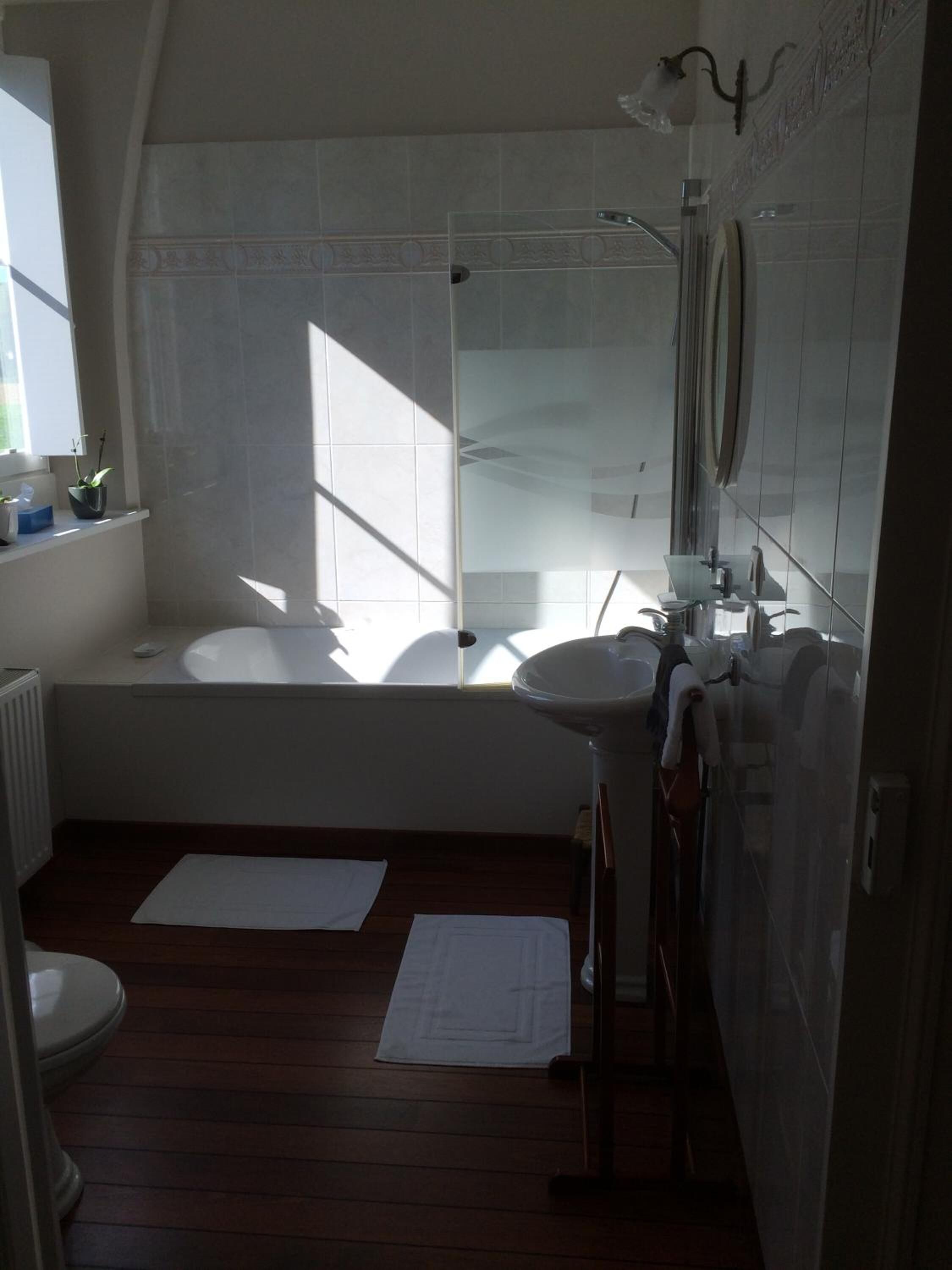 A bathroom with a bathtub, a sink, and a shower. There is a window providing natural light, and the floor is wooden.