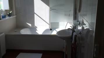 A bathroom with a bathtub, a sink, and a shower. There is a window providing natural light, and the floor is wooden.