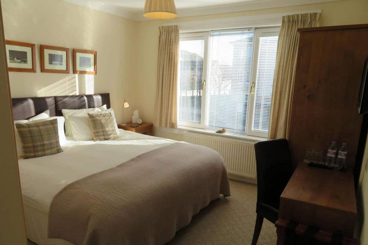 A hotel room with a large bed, a desk, a chair, a window with blinds, and a radiator.