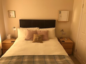 Un lit bien fait, recouvert d’un plaid à carreaux, deux tables de chevet avec des lampes, un tableau encadré au mur et une porte sur le côté droit.