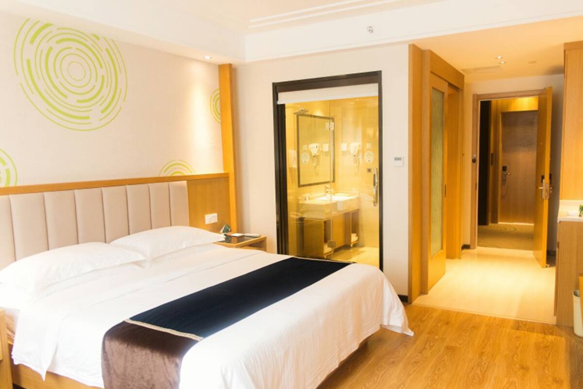 A hotel room with a bed, a nightstand, a bathroom with a glass door, and a wooden floor.