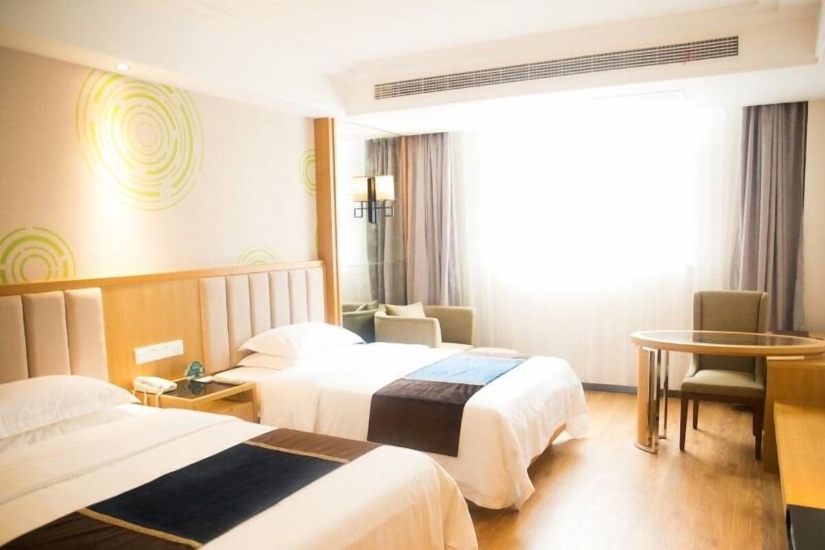 A hotel room with two beds, a desk, and a chair. There is a window with curtains, a wall-mounted phone, and a lamp on the nightstand.