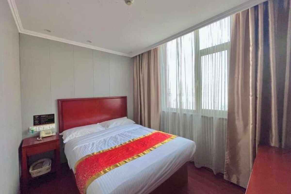 A hotel room with a single bed, a nightstand, a window with curtains, and a view of the outside.