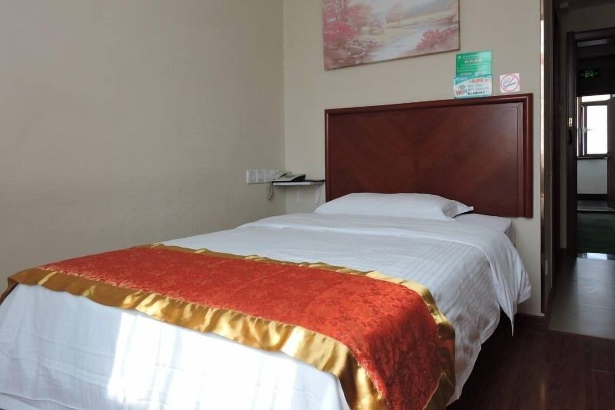 A hotel room with a wooden headboard, a bed with white and red bedding, a painting on the wall, and a door leading to another room.