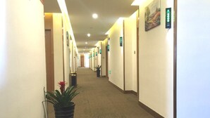 Corredor de hotel com pisos carpetados, plantas em vasos e placas de sinalização nas paredes.