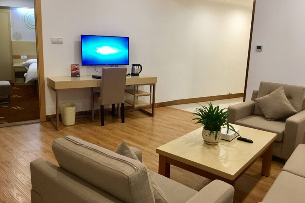 Una sala de estar moderna con piso de madera, un sofá color beige, una mesa de centro con una planta, un televisor sobre un escritorio y una puerta que da a un dormitorio.