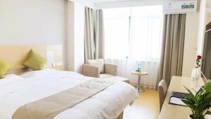 Une chambre d’hôtel avec un grand lit, un bureau, une chaise, une petite table, une plante en pot et une fenêtre avec des rideaux.