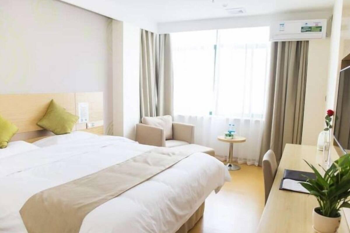 Ein Hotelzimmer mit einem großen Bett, einem Schreibtisch, einem Sessel, einem kleinen Tisch, einer Topfpflanze und einem fenster mit Vorhängen.