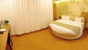 Une chambre d’hôtel avec un lit rond, un bureau avec une chaise et une grande fenêtre avec des rideaux blancs.