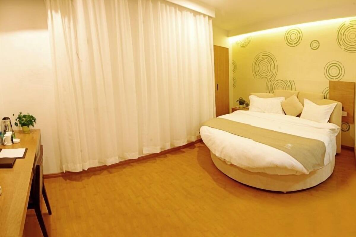 Ein Hotelzimmer mit einem runden Bett, einem Schreibtisch mit Stuhl und einem großen Fenster mit weißen Vorhängen.