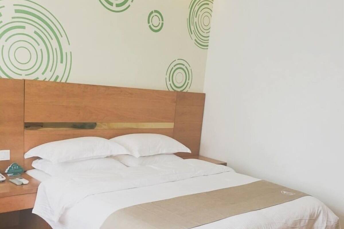 Ein ordentlich bezogenes Bett mit weißen Bettwäsche und einer beigen Tagesdecke, ein hölzernes Kopfende und ein Nachttisch mit Telefon und einem grünen Gegenstand.