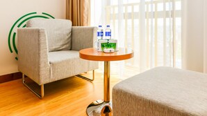 A hotel room with a grey armchair, a round wooden table with a water bottle and a green mug, and a large window with sheer curtains. - GreenTree Inn Wuhu Fangte Four Phase Wanchun Fortune Plaza Hotel (Wuhu)