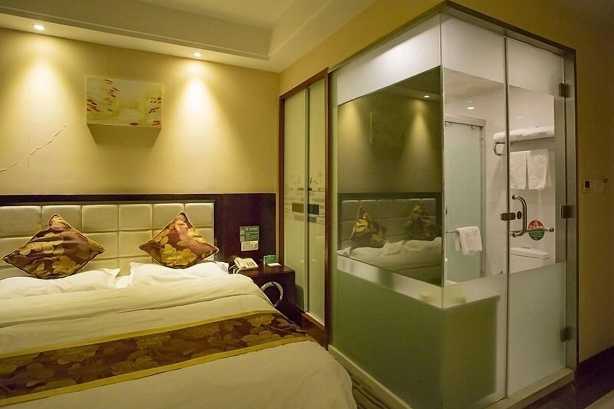 Una habitación de hotel con una cama, una mesita de noche, una zona de ducha con mampara de vidrio y un aplique de pared.