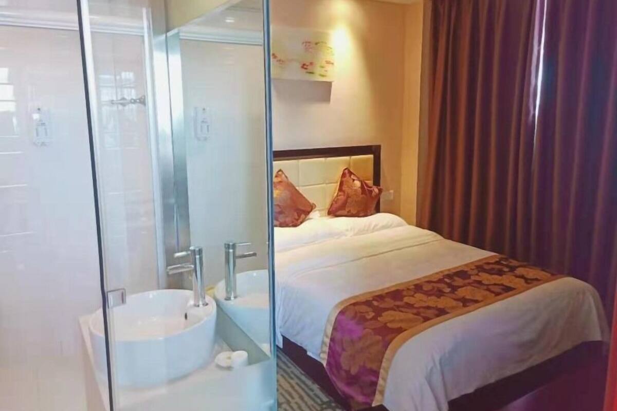 Habitación de hotel con ducha envuelta en vidrio, lavamanos blanco y cama con colcha estampada.