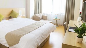 Une chambre d’hôtel avec un grand lit, un bureau avec une chaise, une petite table, une plante en pot et une fenêtre avec des rideaux.