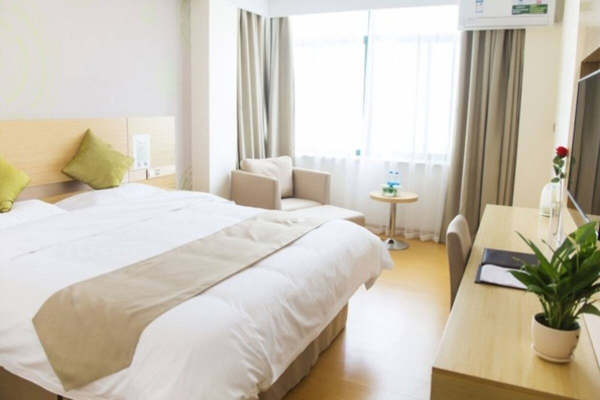 A hotel room with a large bed, a desk with a chair, a small table, a potted plant, and a window with curtains.