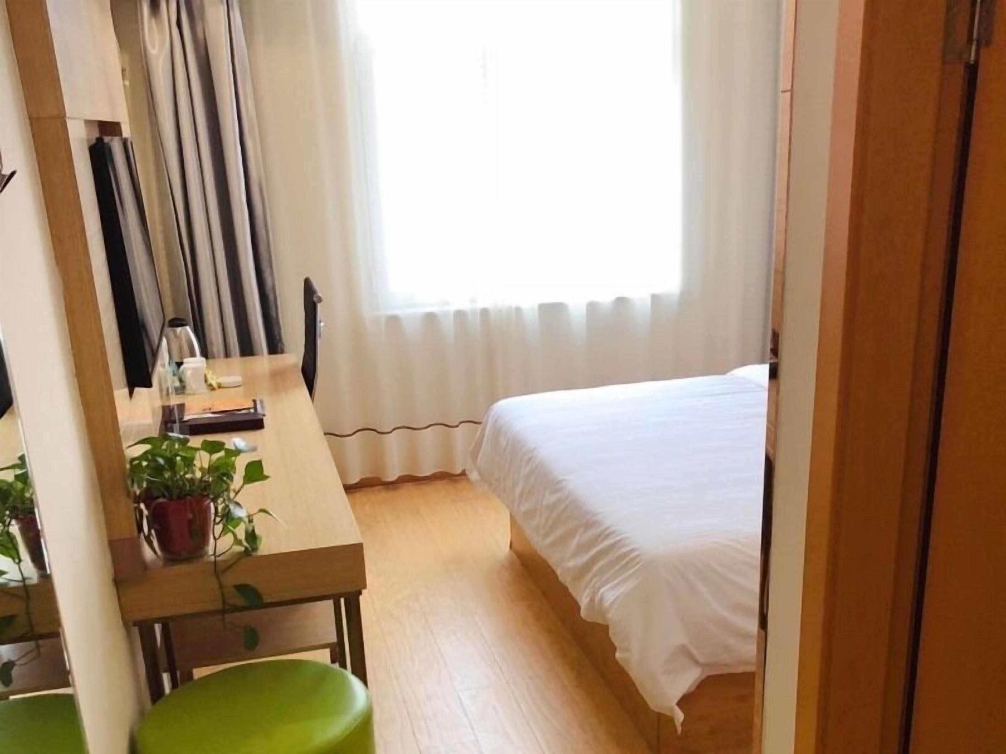 A hotel room with a bed, a desk with a chair, a potted plant, and a window with curtains.