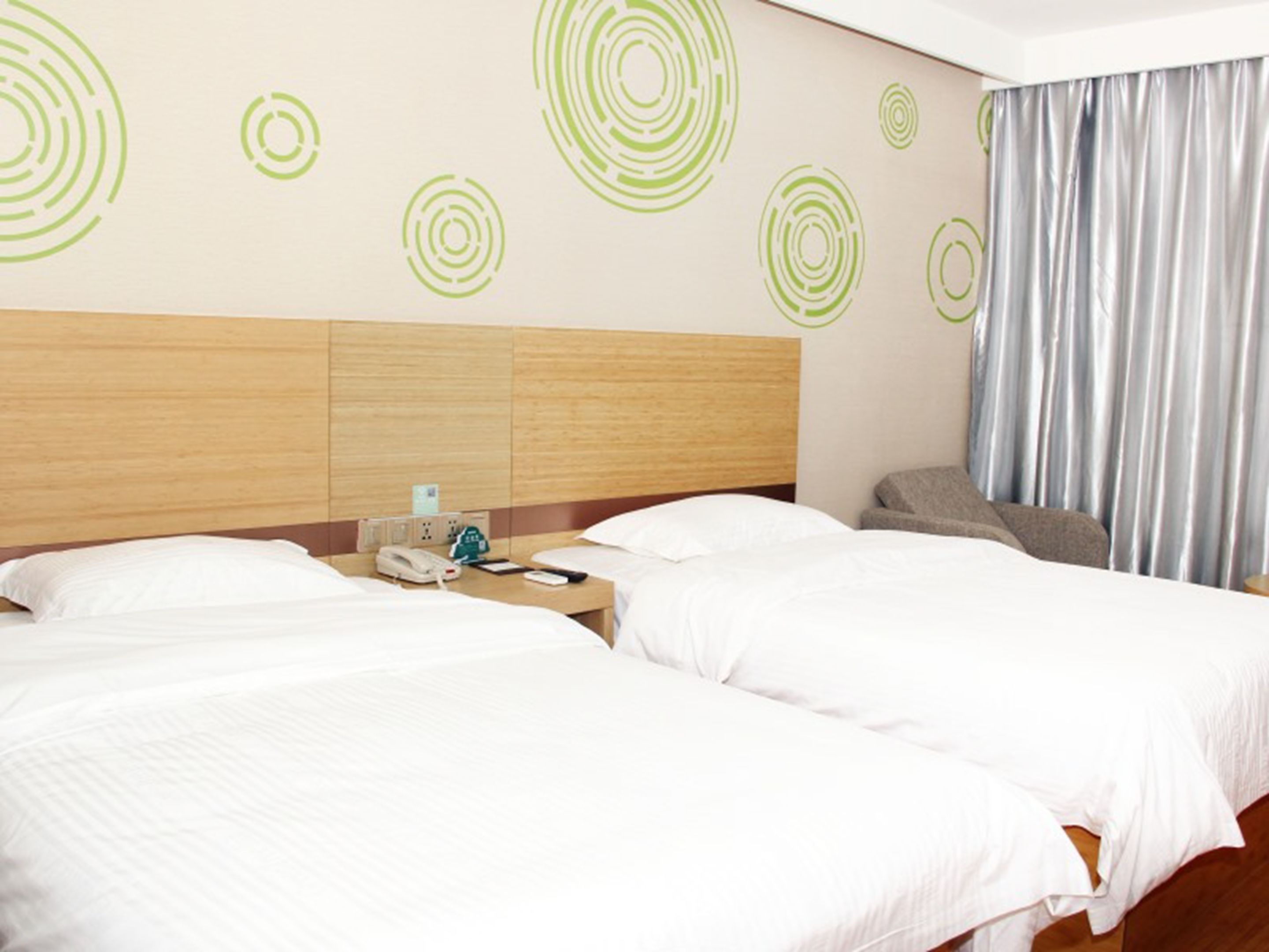 A hotel room with two beds, a wooden headboard, a nightstand with a phone and a clock, and a curtain.