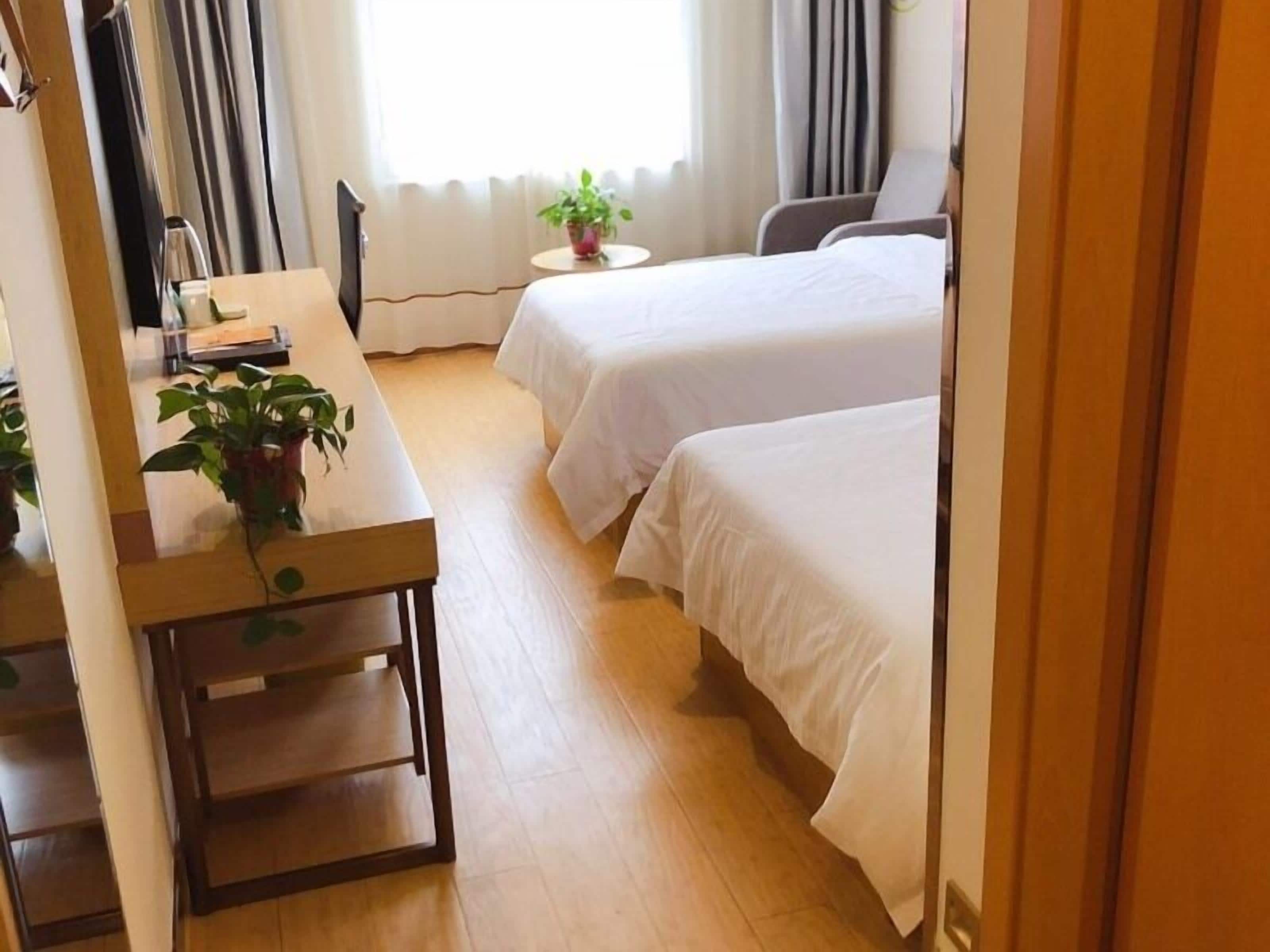 A hotel room with two beds, a desk with a chair, a small table with a plant, and a window with curtains.