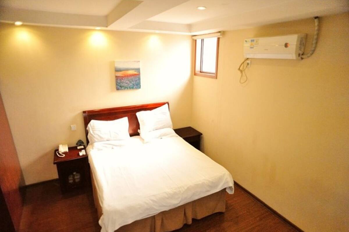 Ein ordentlich bezogenes Bett in einem Hotelzimmer, mit einem hölzernen Kopfteil, einem Nachttisch mit Telefon und einer Klimaanlage an der Wand.