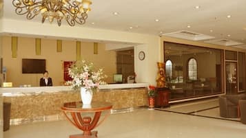 A hotel reception area with a receptionist, a large chandelier, a reception desk, a vase of flowers, and a large window.