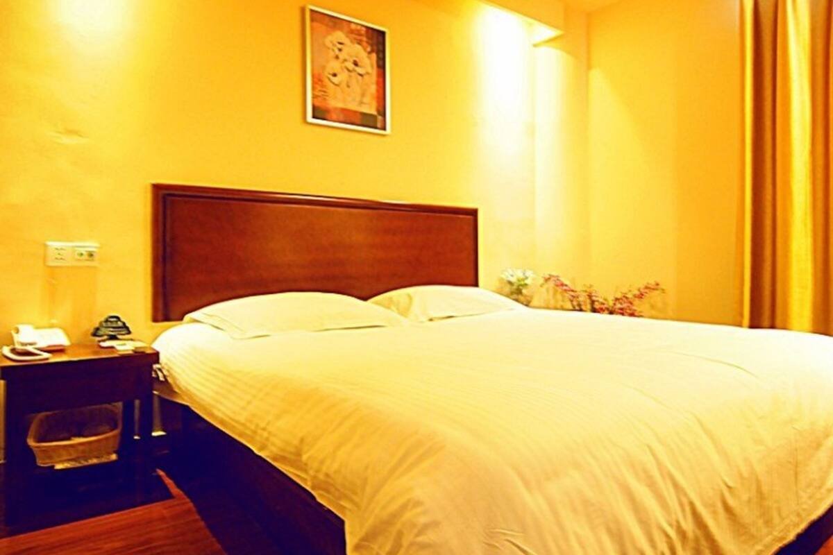 A hotel room with a wooden headboard, a bed with white linens, a nightstand with a phone and a small plant, and a painting on the wall.
