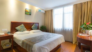 A hotel room with a wooden bed, a nightstand, a desk with a chair, a potted plant, and a window with curtains.