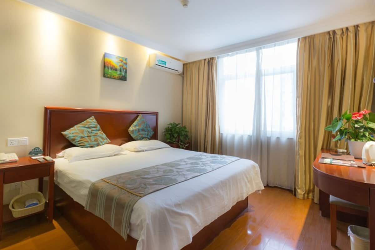 Une chambre d’hôtel avec un lit en bois, une table de chevet, un bureau avec une chaise, une plante en pot et une fenêtre avec des rideaux.