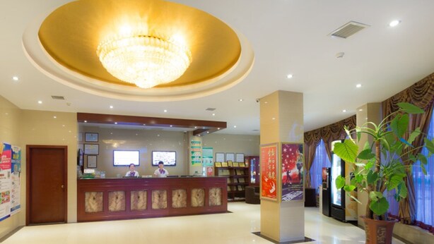 A hotel lobby with a reception desk, a large chandelier, a potted plant, and a poster on the wall.